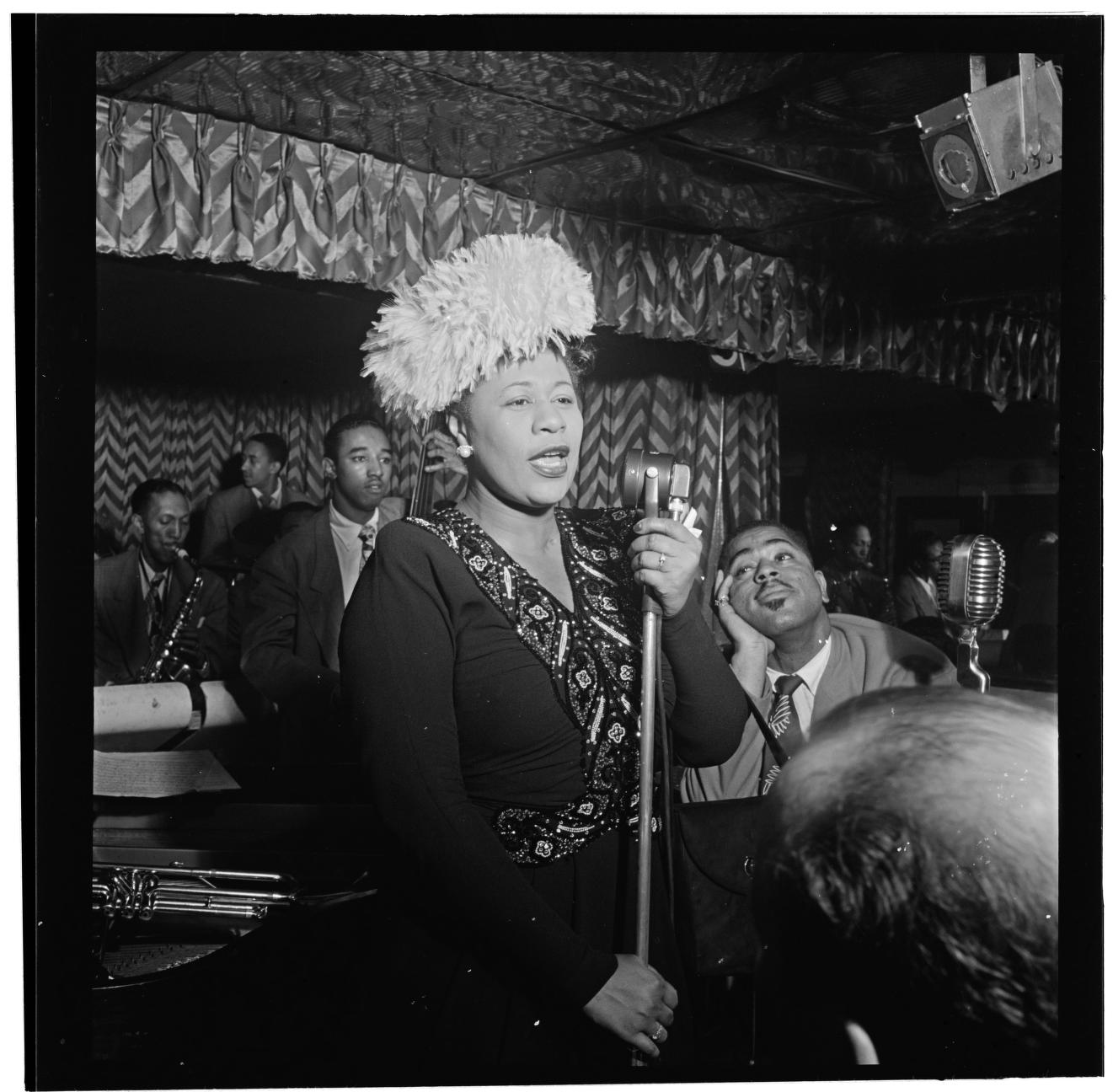 Ella Fitzgerald canta ante un micrófono de pie en un club lleno, con un tocado de plumas y un vestido bordado. A su derecha, Dizzy Gillespie la mira con la barbilla apoyada en la mano; detrás hay otros músicos, un clarinete y un piano.